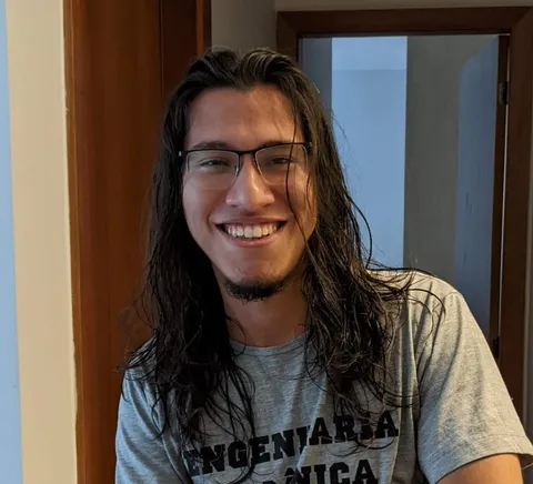 Casual phone photo of a smiling man with long hair and glasses in a hallway