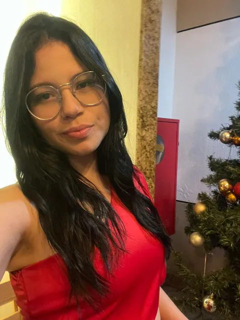 Phone selfie of a woman with round glasses and a red top beside a Christmas tree